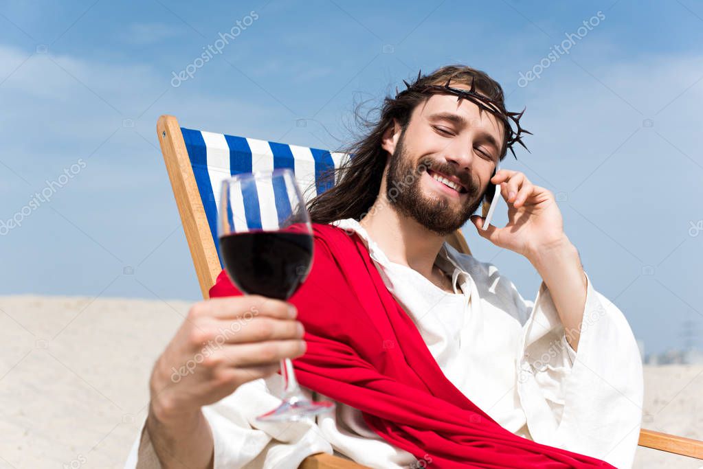 happy Jesus resting on sun lounger with glass of wine and talking by smartphone in desert