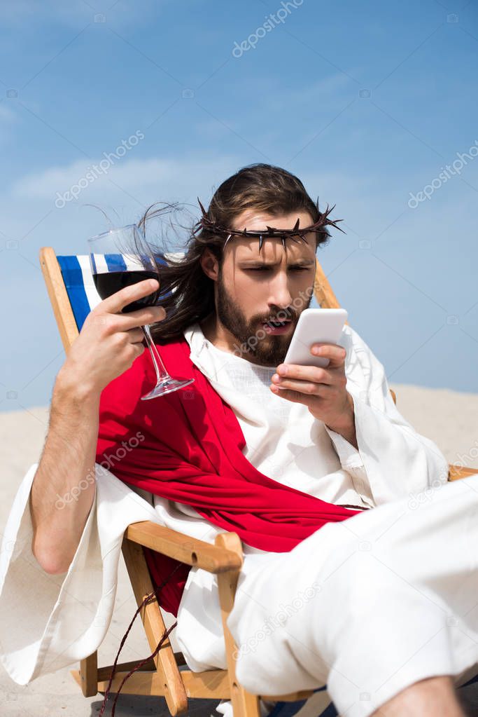 surprised Jesus resting on sun lounger with glass of wine and looking at smartphone in desert