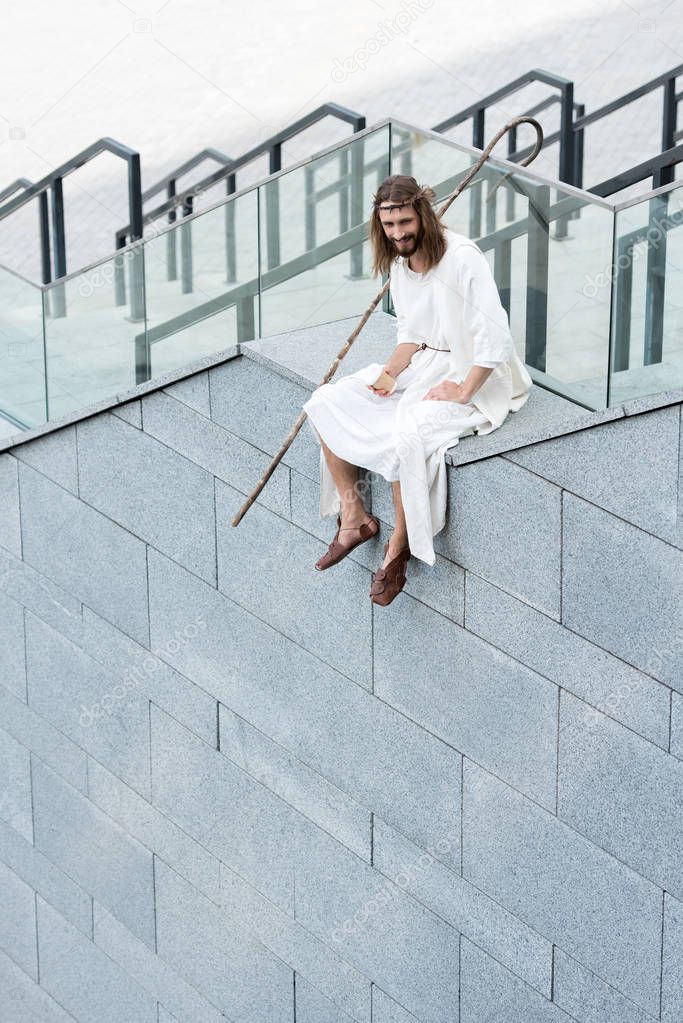 smiling Jesus in robe and crown of thorns sitting on staircase side, holding disposable coffee cup and looking down