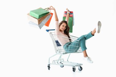 excited girl with shopping bags sitting in shopping cart, isolated on white