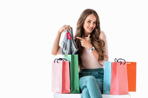 happy young woman with shopping bags pointing at clothes with sale tag, isolated on white