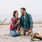 Женщина кормит мужа зефиром у костра на песчаном пляже Женщина кормит мужа зефиром у костра на песчаном пляже