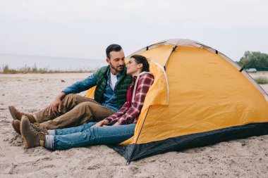 kumlu plajda kamp çadır oturan çift yan görünüm