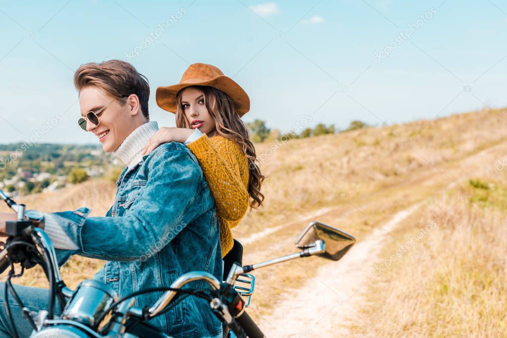 Girlfriend looking at camera while boyfriend sitting on vintage motorbike on rural meadow