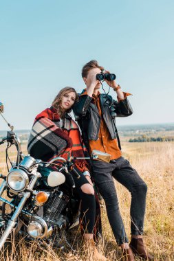 young boyfriend looking through binoculars near girlfriend sitting on motorbike  clipart