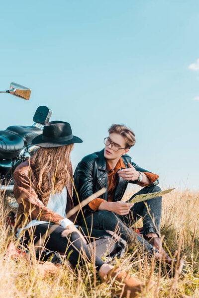 young boyfriend in glasses holding map and looking at beautiful girlfriend sitting on meadow 