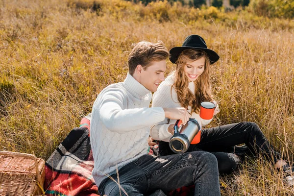 年轻的微笑的夫妇倾吐热的饮料从热水瓶在野餐在农村草甸 — 免费的图库照片