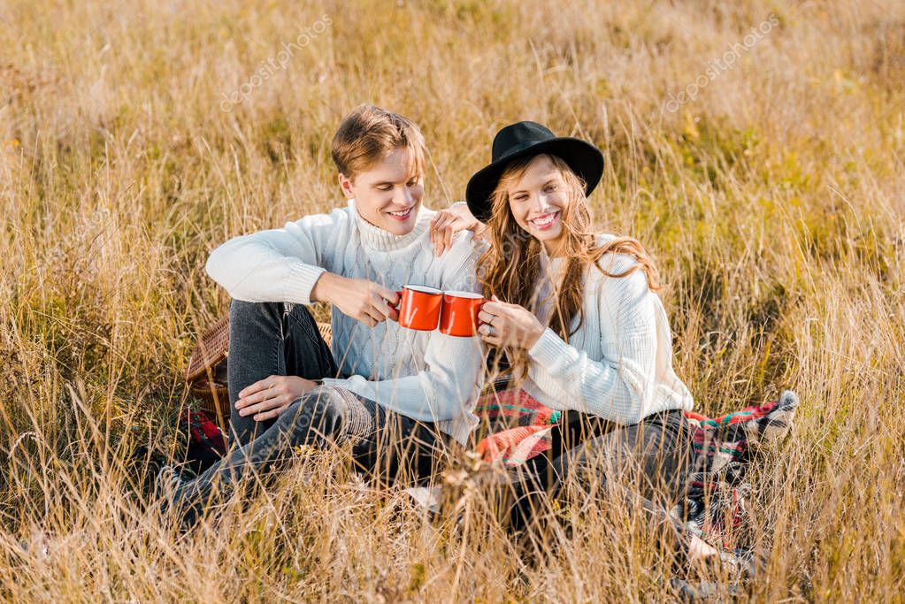 Young couple clinking with cups and sitting on plaid on rural meadow