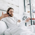 Hombre de negocios barbudo enojado con el pelo largo leyendo el periódico y el gesto a mano en la cama durante el horario de la mañana en casa