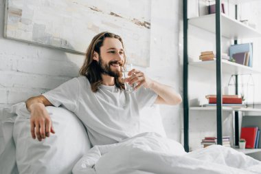 smiling bearded man with long hair drinking water in bedroom at home  clipart