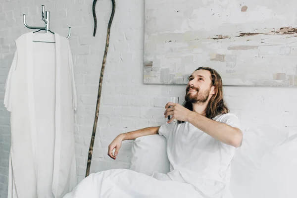 happy Jesus looking up and holding glass of water in bed during morning time at home