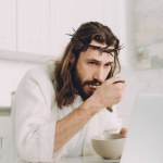Selective focus of Jesus eating corn flakes on breakfast at table with laptop in kitchen at home