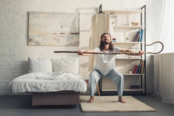 smiling Jesus in crown of thorns doing morning workout with wooden staff in bedroom at home