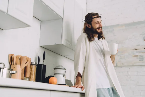 low angle view of smiling Jesus in crown of thorns standing with cup of coffee in kitchen at home