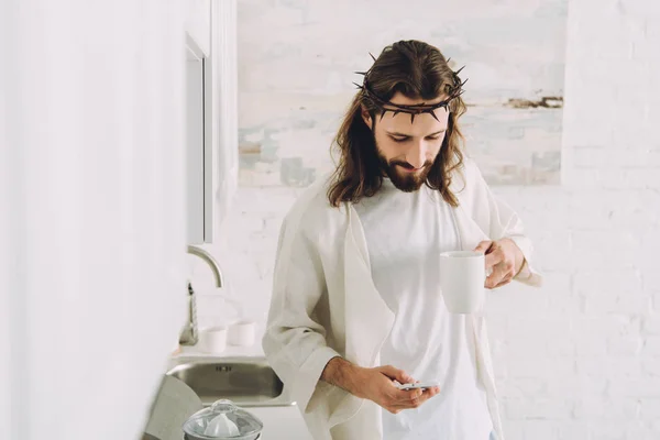 Счастливый Иисус Терновом Венце Стоит Чашкой Кофе Пользуется Смартфоном Кухне — Бесплатное стоковое фото