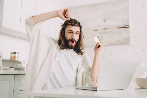 excited Jesus with credit card gesturing by hands and doing online shopping at table with laptop in kitchen at home