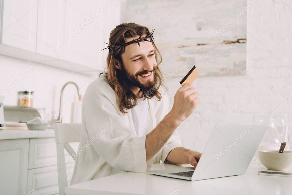 smiling Jesus holding credit card and doing online shopping at table with laptop in kitchen at home