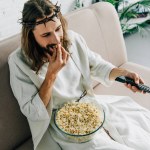 Vysoký úhel pohled na Ježíše v trnová koruna, sledování televize a jíst popcorn na gauči doma