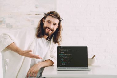 happy Jesus holding cup of coffee and pointing at laptop with html code in kitchen at home clipart
