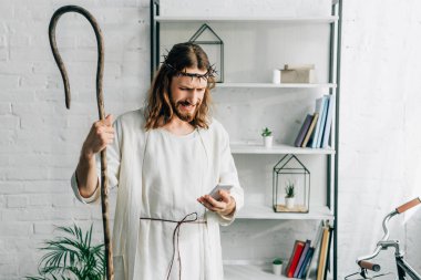 emotional Jesus in robe with wooden staff using smartphone at home clipart