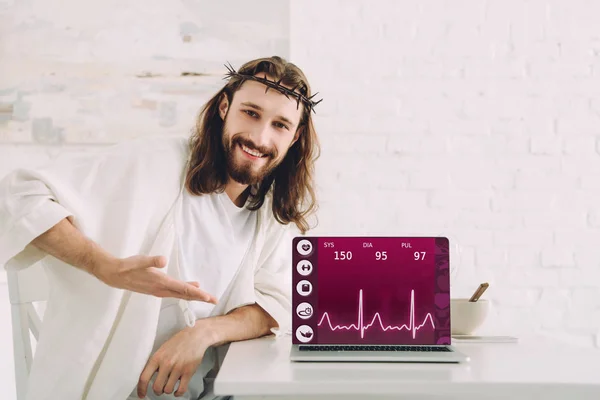 快乐的耶稣在荆棘王冠指向笔记本电脑在屏幕上的医疗应用在家里的厨房 — 图库照片