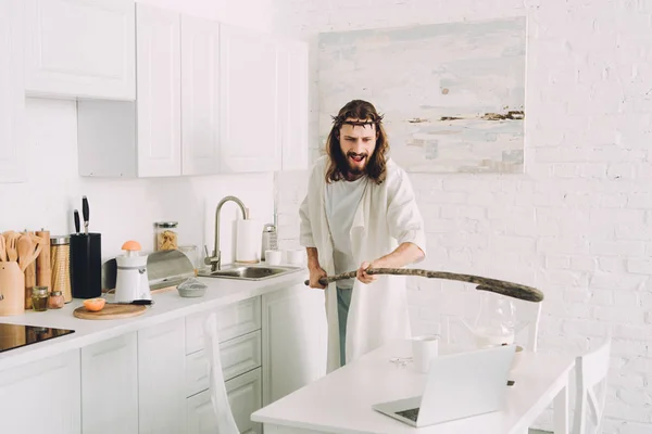 aggressive Jesus hitting laptop by wooden staff in kitchen at home