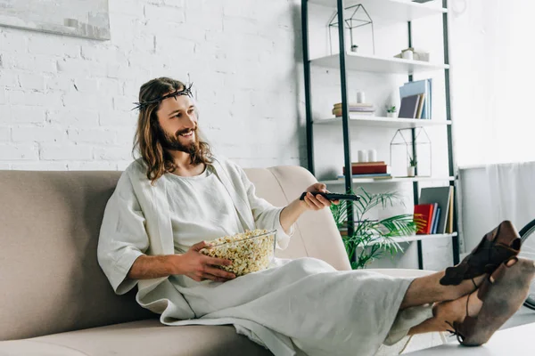 happy Jesus in crown of thorns watching tv and sitting with bowl of popcorn on sofa at home