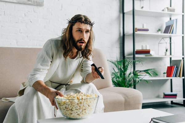 shocked Jesus in crown of thorns watching tv and sitting with bowl of popcorn on sofa at home