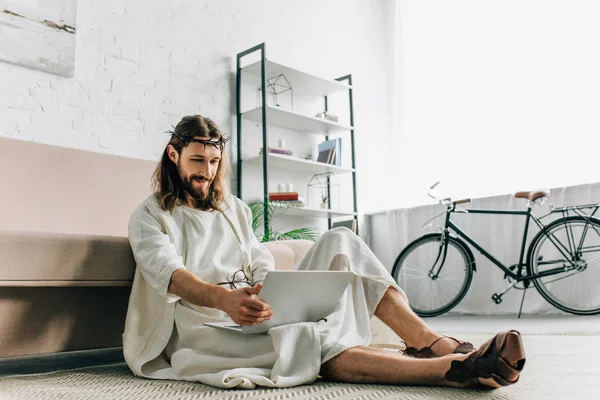 smiling Jesus sitting on floor and using laptop near sofa at home