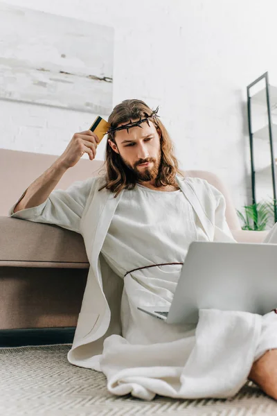 pensive Jesus sitting on floor with credit card and doing online shopping on laptop near sofa at home