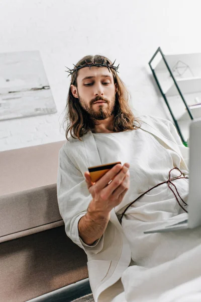 low angle view of pensive Jesus looking at credit card and doing online shopping on laptop near sofa at home