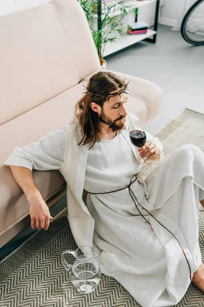 high angle view of Jesus in crowns of thorns and robe sitting on floor near jug of water and drinking wine from glass at home