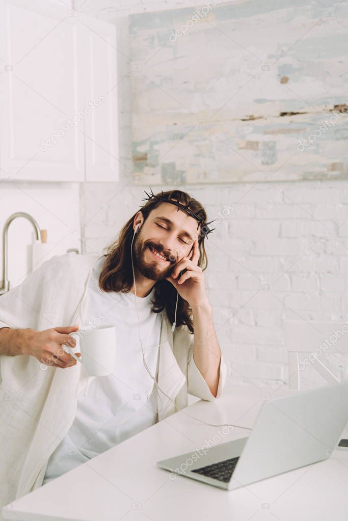 cheerful Jesus in earphones listening music and holding cup of coffee at table with laptop in kitchen at home
