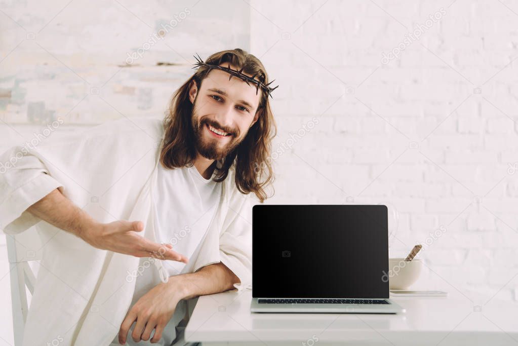 smiling Jesus in crown of thorns pointing at laptop with blank screen in kitchen at home