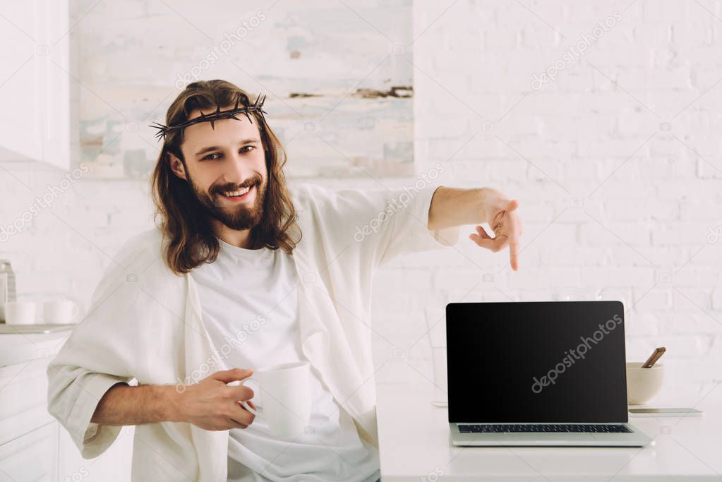 happy Jesus holding cup of coffee and pointing at laptop with blank screen in kitchen at home