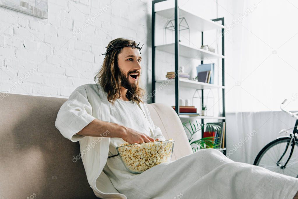 cheerful Jesus in crown of thorns watching tv and sitting with bowl of popcorn on sofa at home