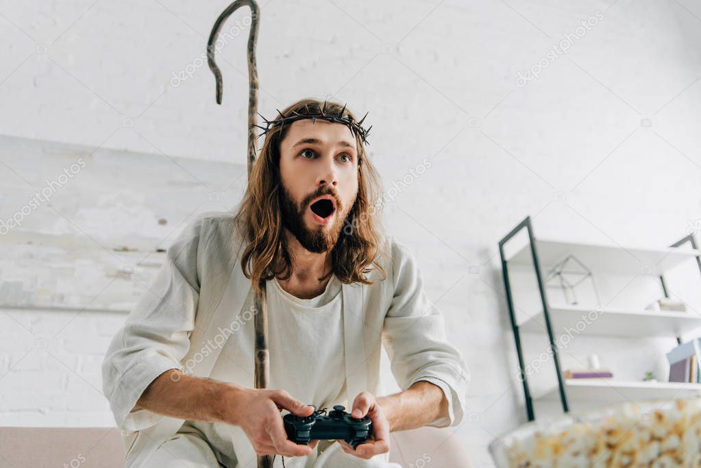 shocked Jesus with wooden staff playing video game by joystick on sofa at home