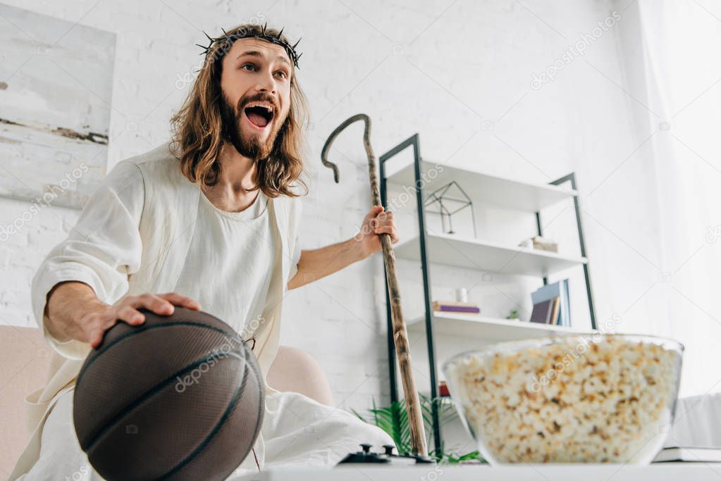 low angle view of excited Jesus with wooden staff holding ball and watching basketball match on sofa at home