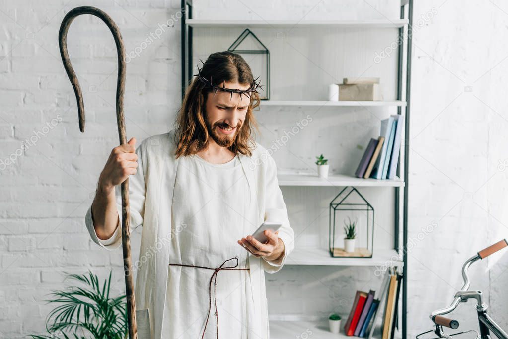 emotional Jesus in robe with wooden staff using smartphone at home