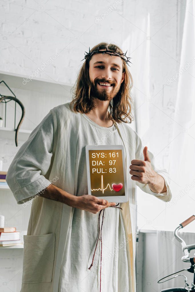 cheerful Jesus in crown of thorns doing thumb gesture and showing digital tablet with medical application at home