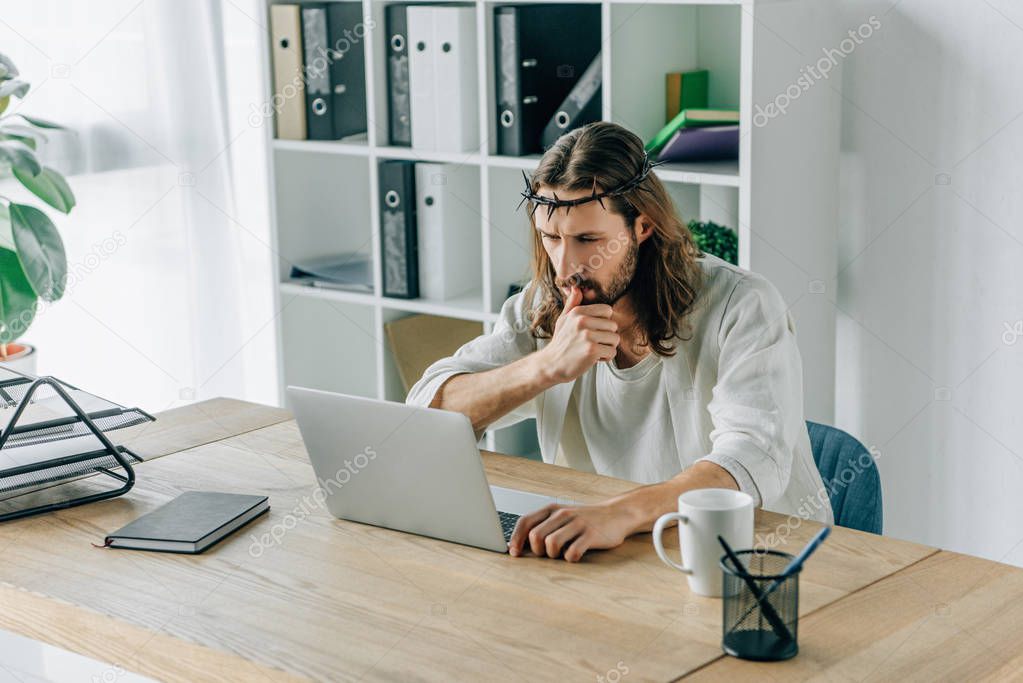 pensive Jesus in crown of thorns using laptop at table in modern office