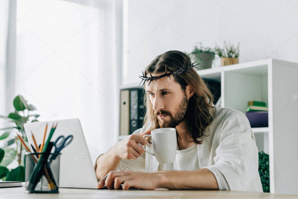 confident Jesus in crown of thorns with coffee cup using laptop at table in modern office