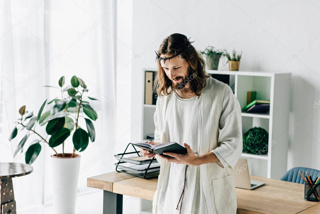 smiling Jesus in crown of thorns and robe reading textbook near working table in modern office 