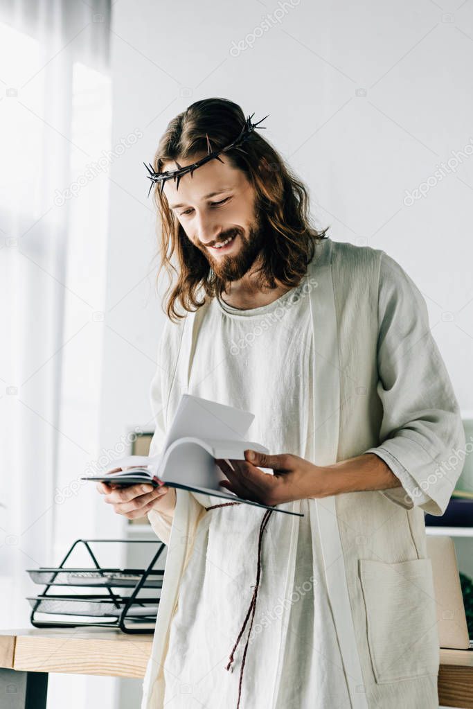 selective focus of happy Jesus in crown of thorns and robe reading textbook near working table in modern office 