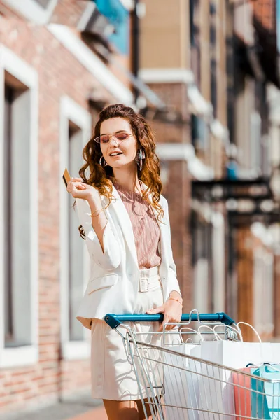 Glückliche Junge Frau Mit Einkaufswagen Voller Papiertüten Mit Kreditkarte — kostenloses Stockfoto