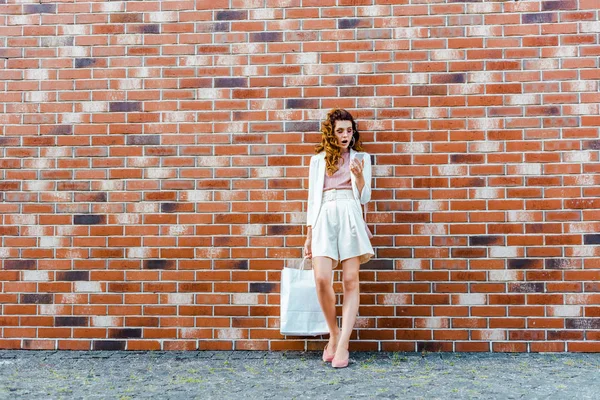 Geschokt Jonge Vrouw Met Boodschappentassen Gebruikend Smartphone Voor Bakstenen Muur — Stockfoto