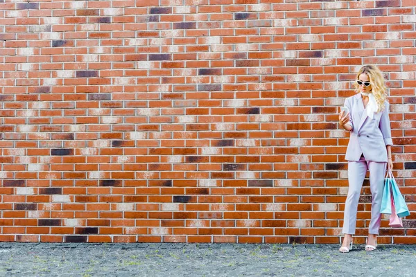 Stijlvolle Jongedame Met Boodschappentassen Gebruikend Smartphone Terwijl Voorkant Van Bakstenen — Stockfoto