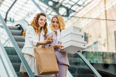 Alışveriş torbaları ve smartphone vasıl alışveriş merkezinde yürüyen merdiven üzerinde arıyorsunuz kutuları olan güzel genç kadın