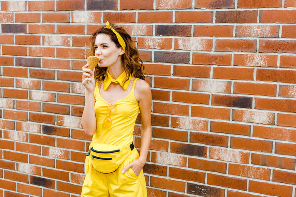 stylish young woman in yellow clothes eating ice cream in front of brick wall