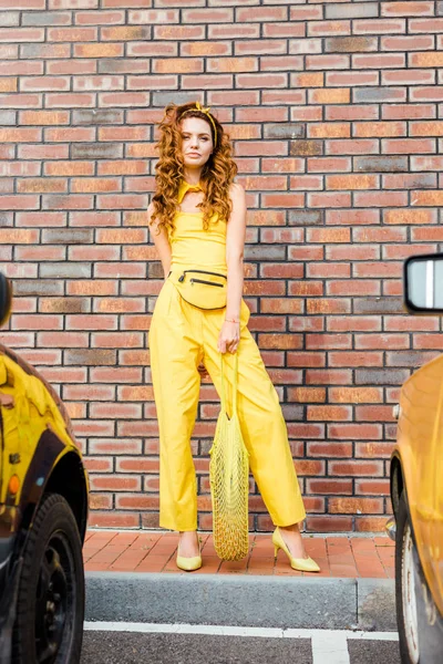 beautiful young woman in yellow clothes with pineapple in string bag standing at parking in front of brick wall